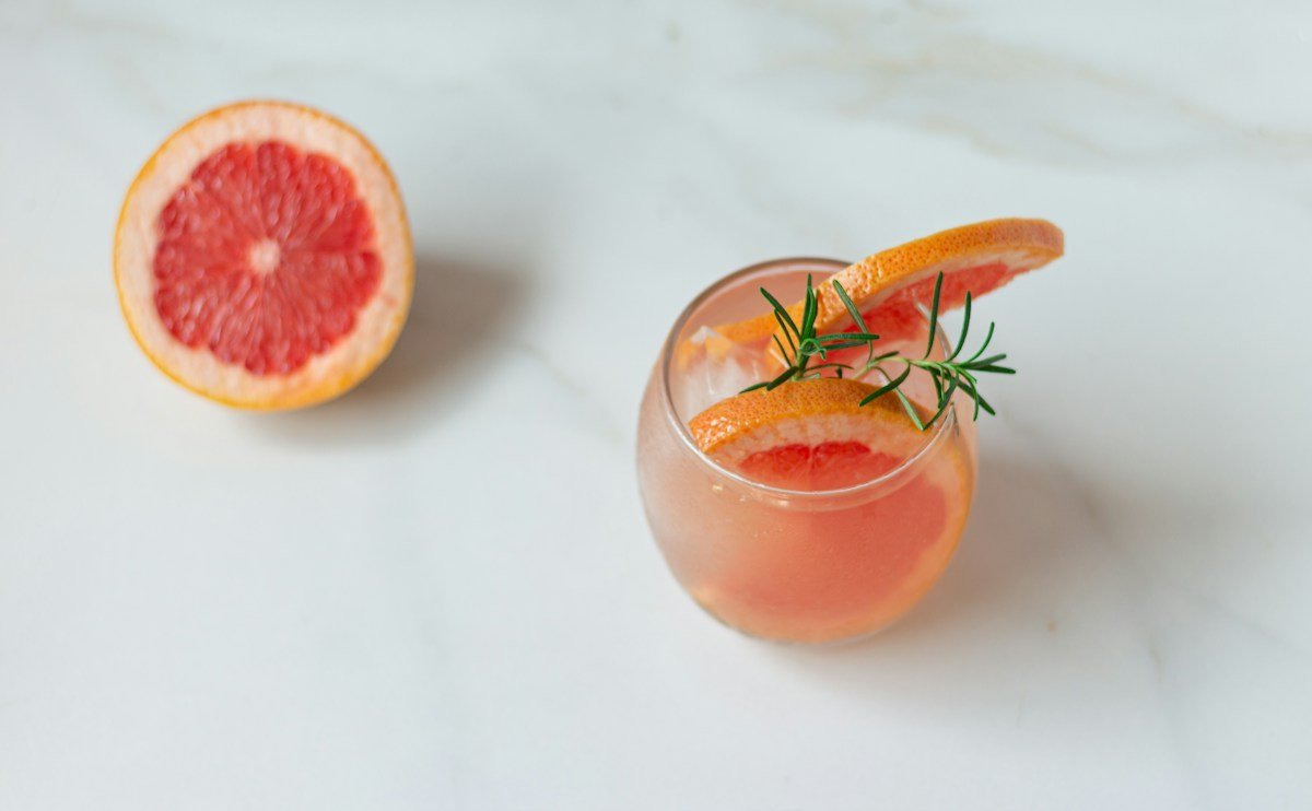 Ein Glas mit Grapefruitgetränk und Rosmarin, daneben eine halbe Grapefruit